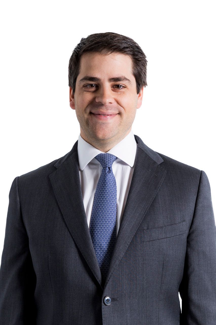 Homem de terno escuro e gravata azul, em pé contra um fundo branco e sorrindo para a câmera.
