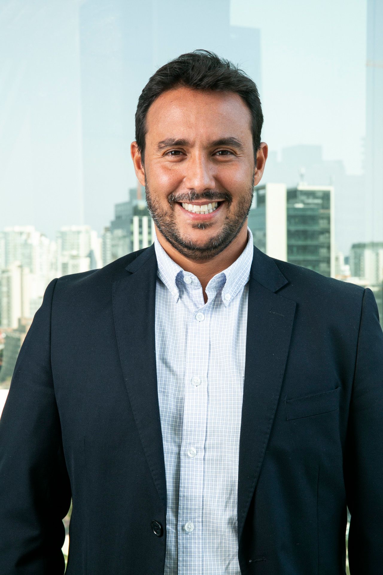Homem sorridente, vestindo blazer azul-marinho e camisa branca, em pé em um escritório com janelas da cidade ao fundo.