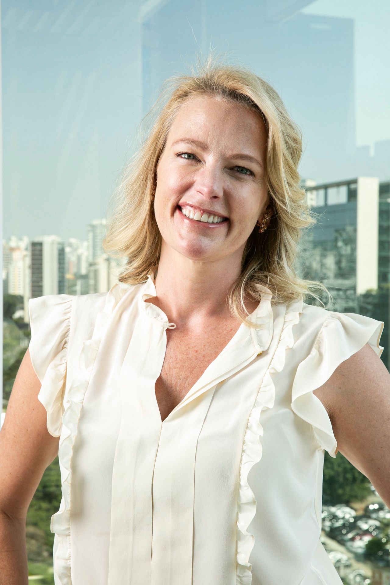 Mulher sorridente de blusa branca em pé diante de uma vista panorâmica da cidade e janelas de vidro.
