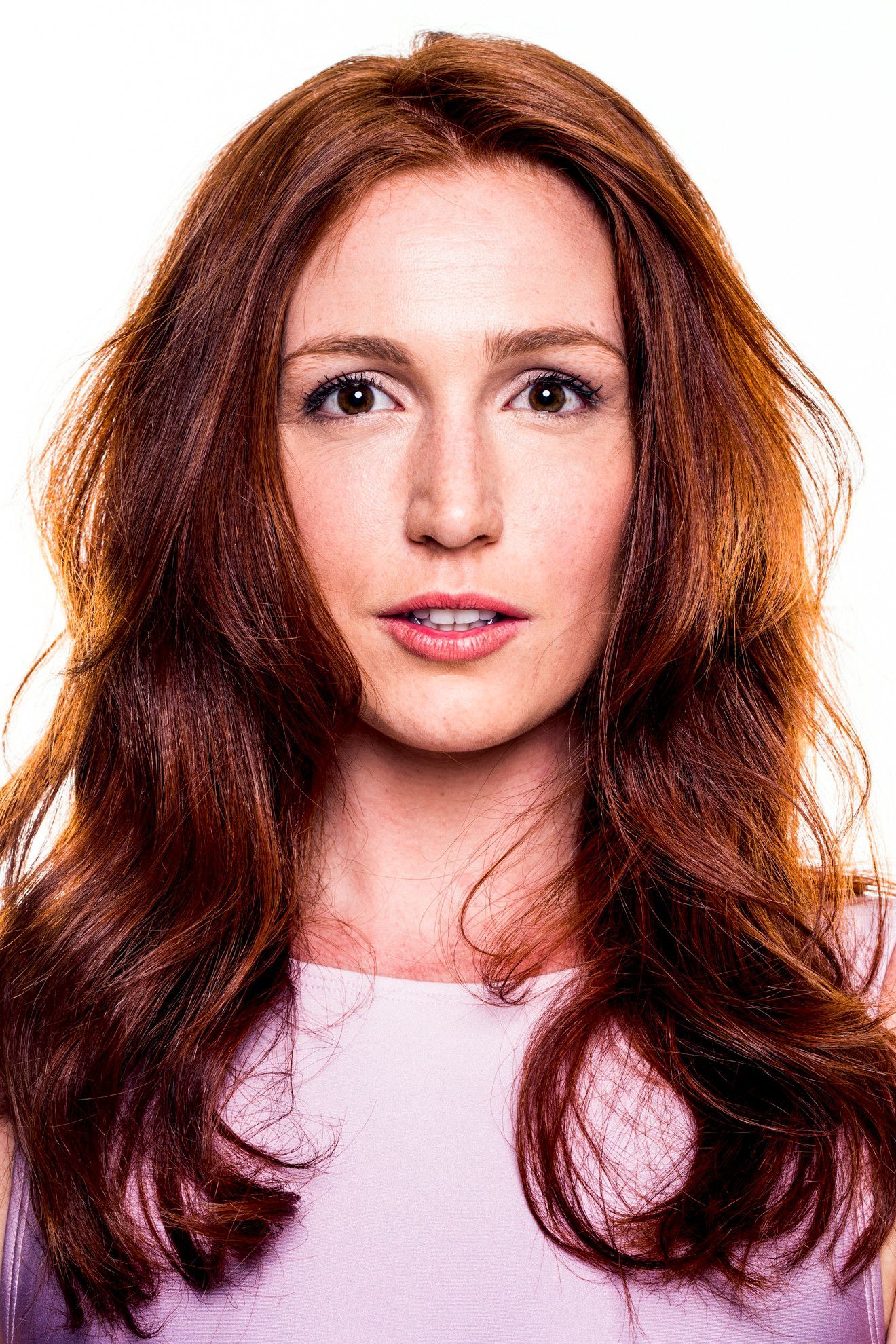 Retrato de uma mulher ruiva de cabelos longos, vestindo uma blusa rosa claro, de frente para um fundo branco.