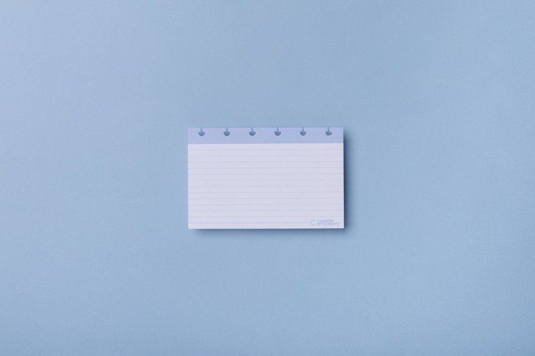 Pequeno cartão de anotações branco com linhas e abas azuis sobre fundo azul claro.
