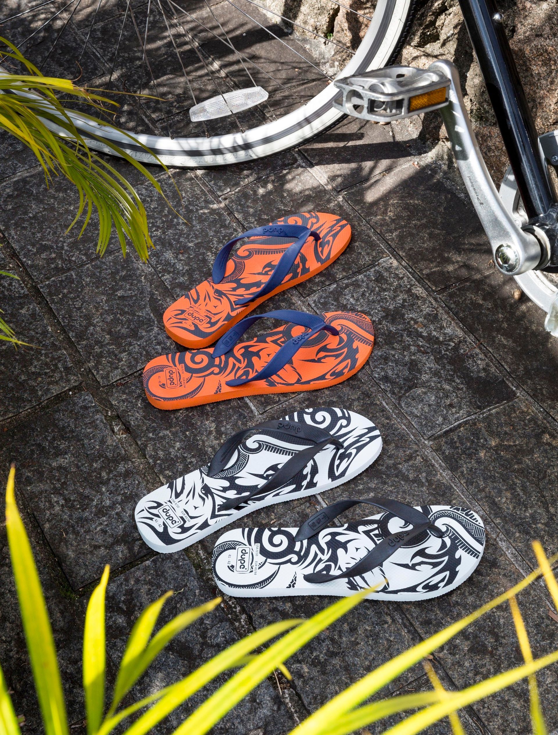 Chinelo laranja e preto empilhado sobre chinelo preto e branco em uma trilha rochosa com rodas de bicicleta por perto.