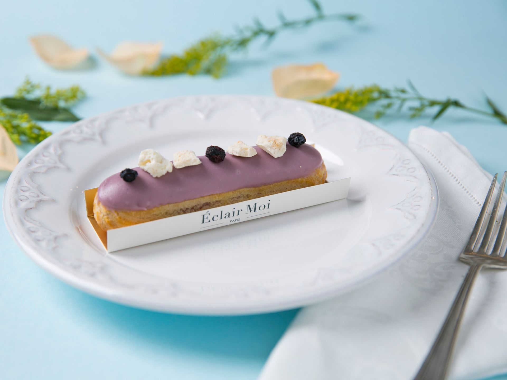 Éclair com cobertura roxa em um prato branco com decoração, ao lado de um garfo e guardanapo em uma mesa azul.