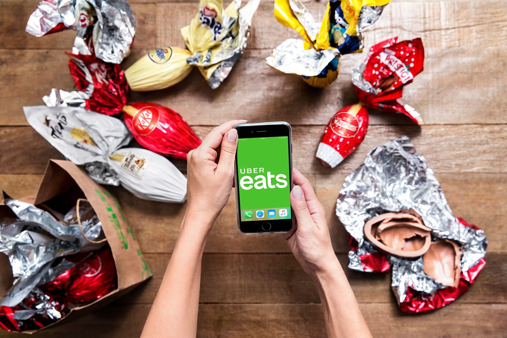 Mãos segurando um celular com o logotipo do Uber Eats, cercadas por embalagens vazias de fast-food sobre uma mesa de madeira.