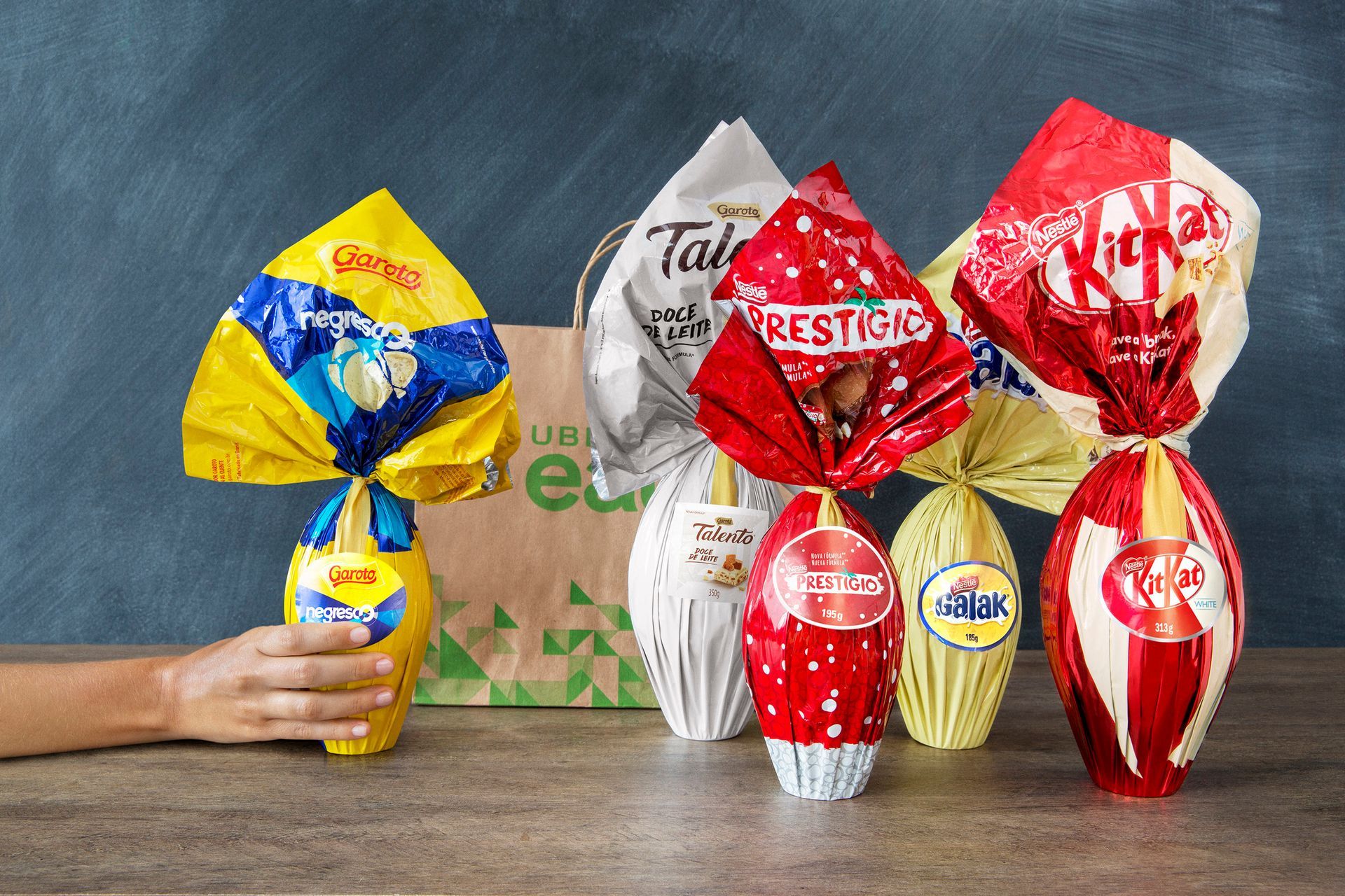 Ovos de Páscoa de chocolate coloridos e coelhinhos embrulhados em uma mesa, com uma mão segurando um deles à esquerda.