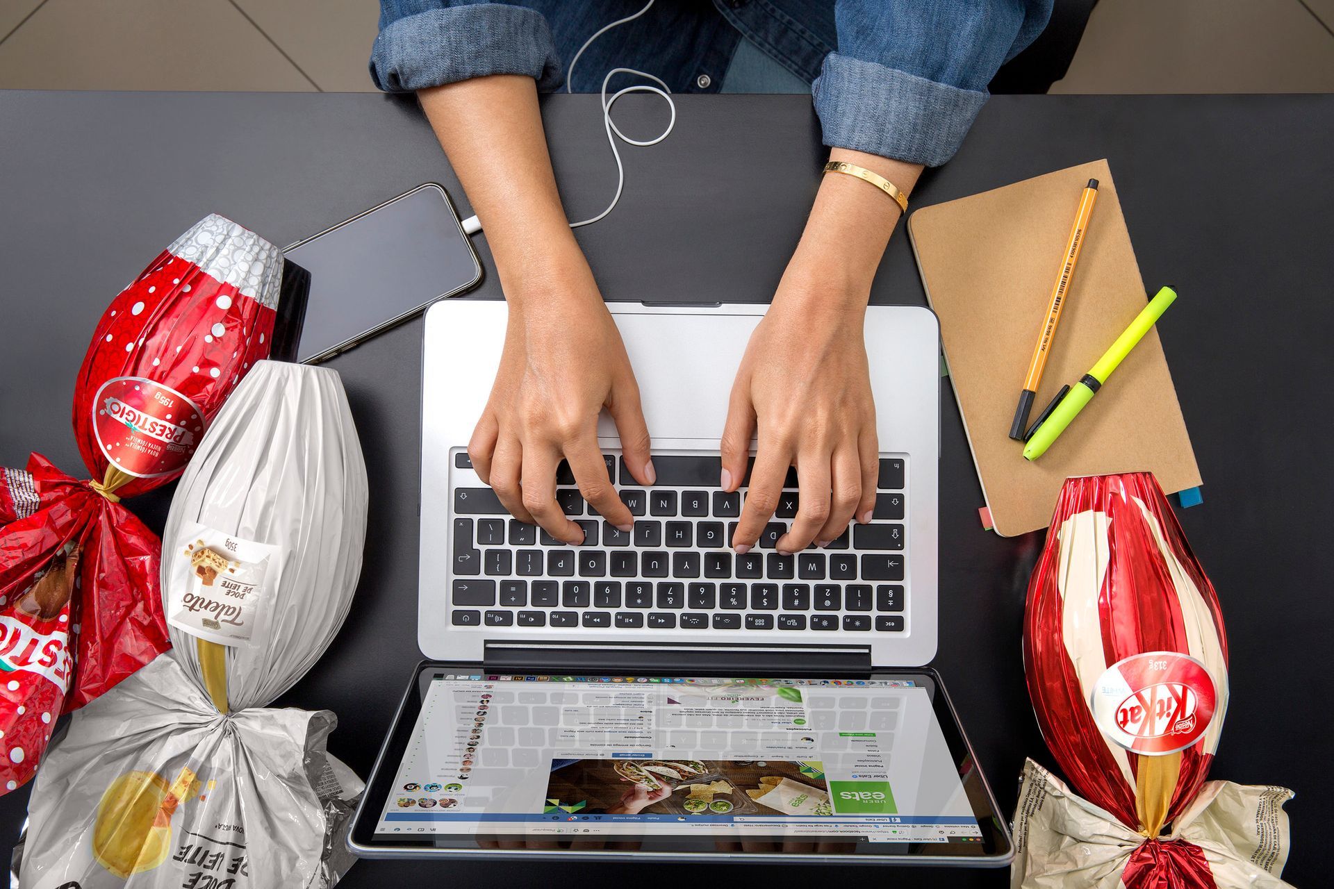 Mãos digitando em um laptop em uma mesa com pacotes de presentes embrulhados, um telefone e lápis.