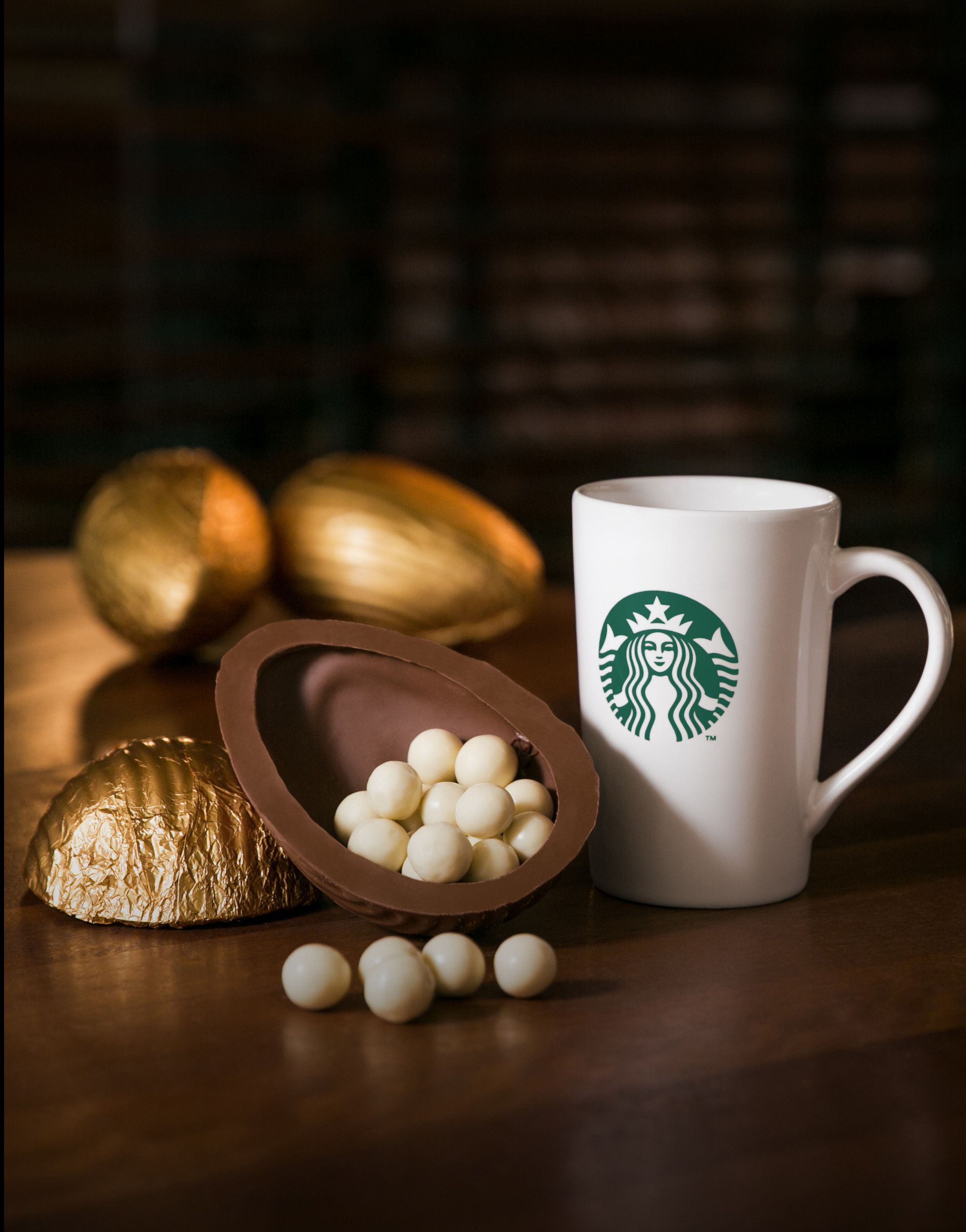 Caneca da Starbucks ao lado de ovos de chocolate e uma casca quebrada sobre uma mesa escura.