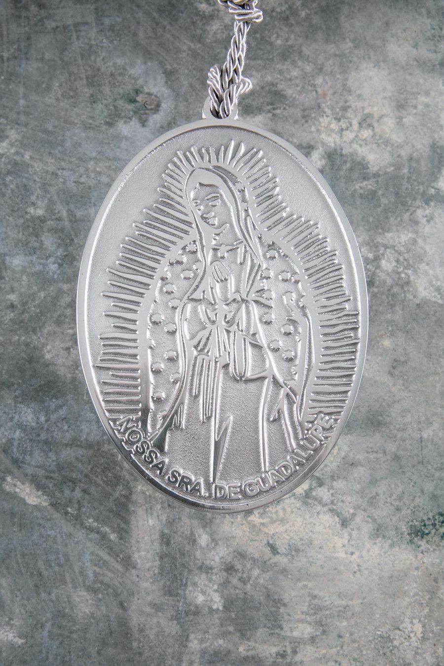 Pingente religioso oval prateado em corrente sobre fundo cinza.