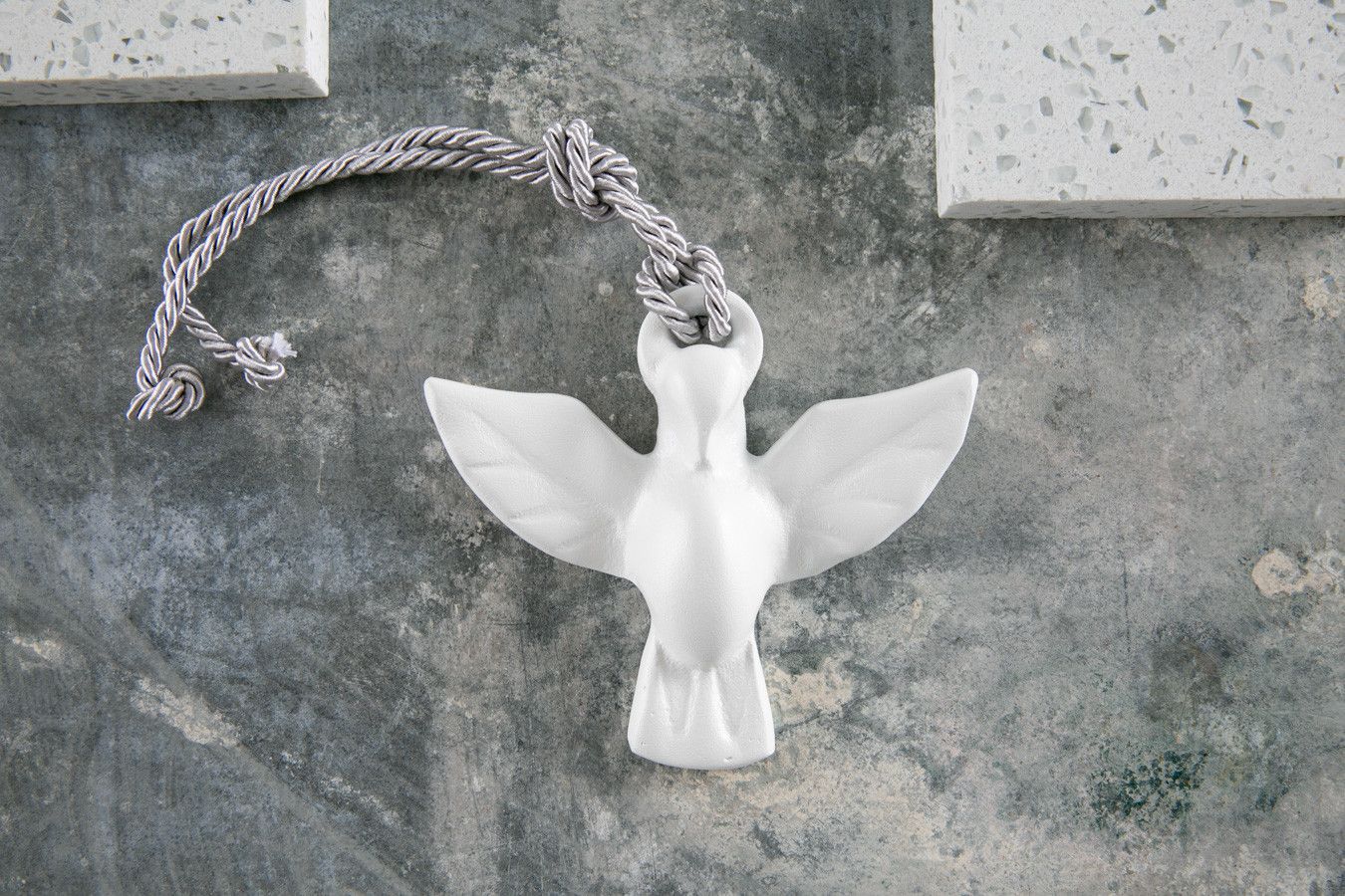 Anjo decorativo de cerâmica branca com asas abertas, pendurado por um cordão trançado em uma superfície texturizada cinza.