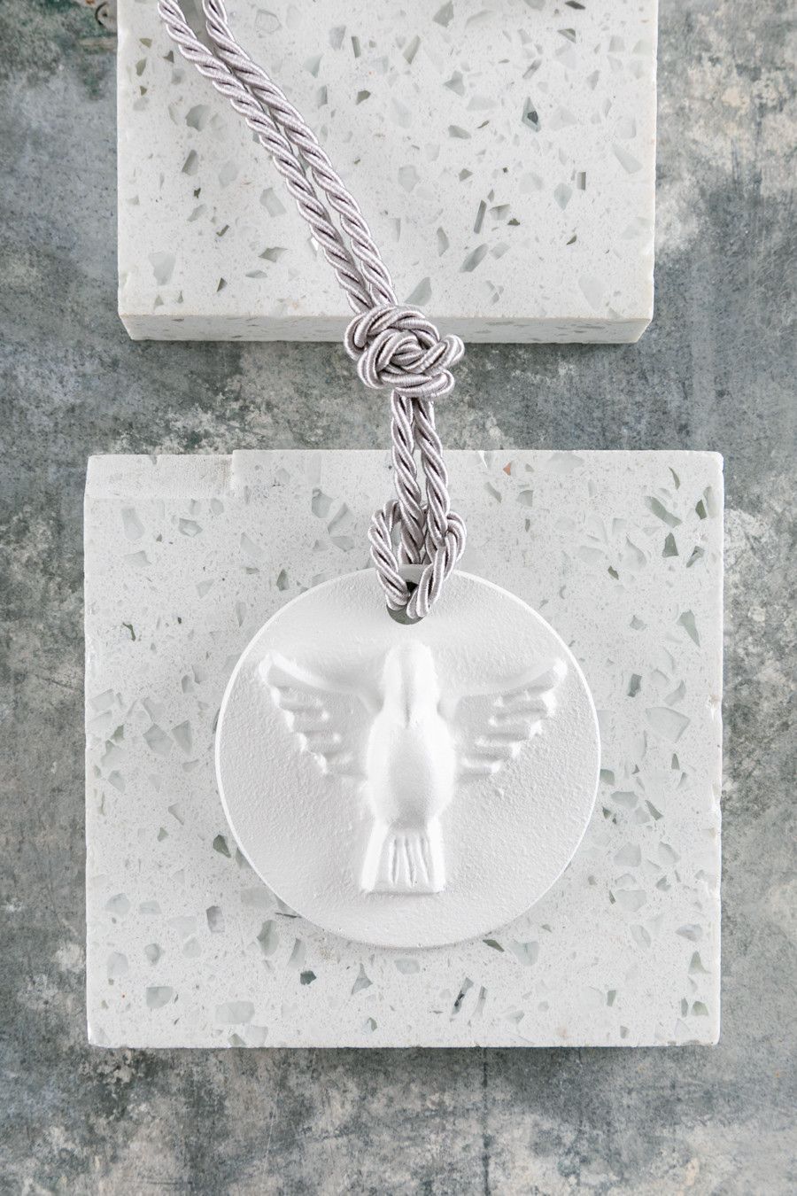 Pingente medalhão branco com um anjo em um cordão trançado sobre um fundo de pedra cinza.