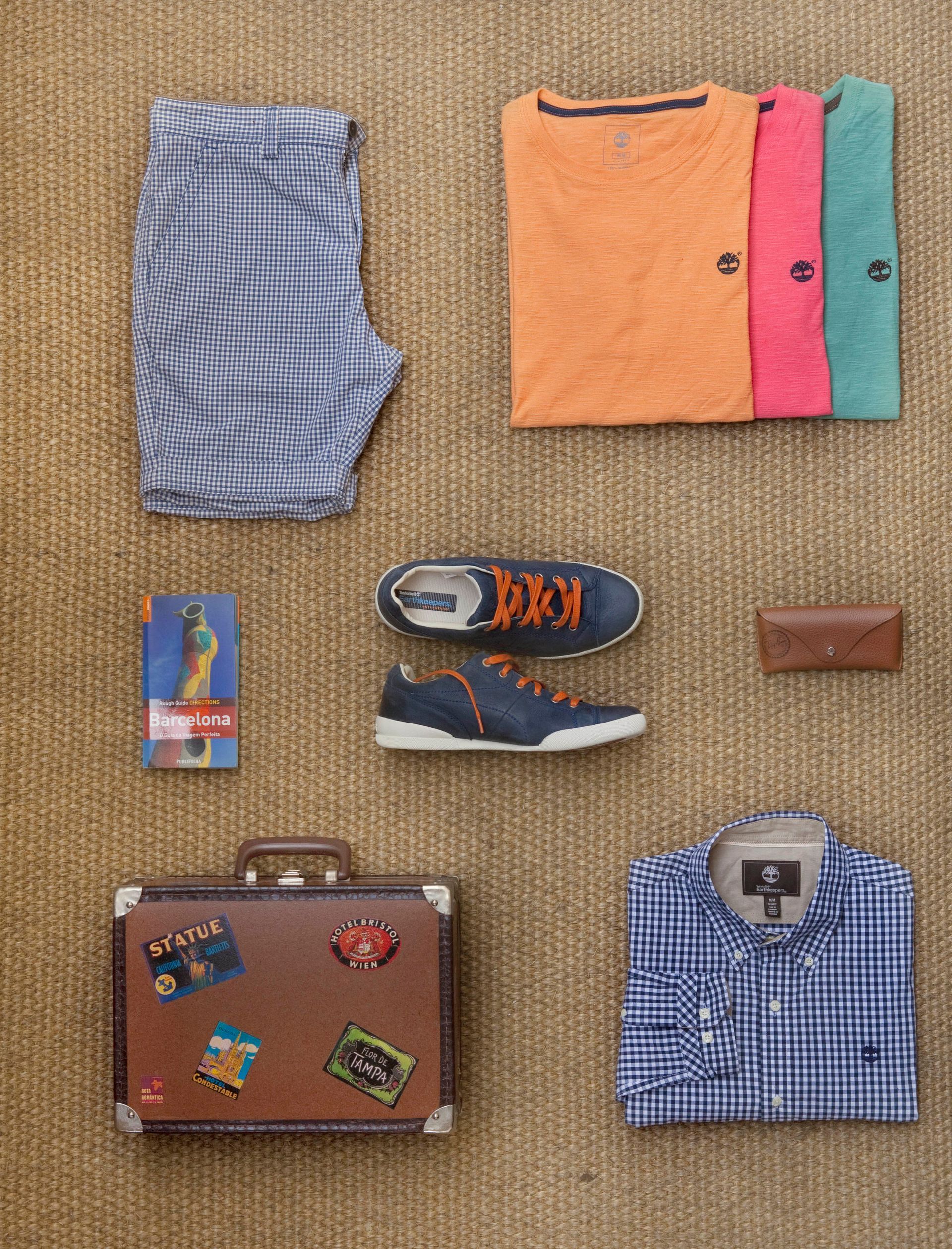 Itens essenciais de viagem dispostos sobre um saco de juta: roupas dobradas, sapatos, cinto, carteira, livros, pasta e camisa.