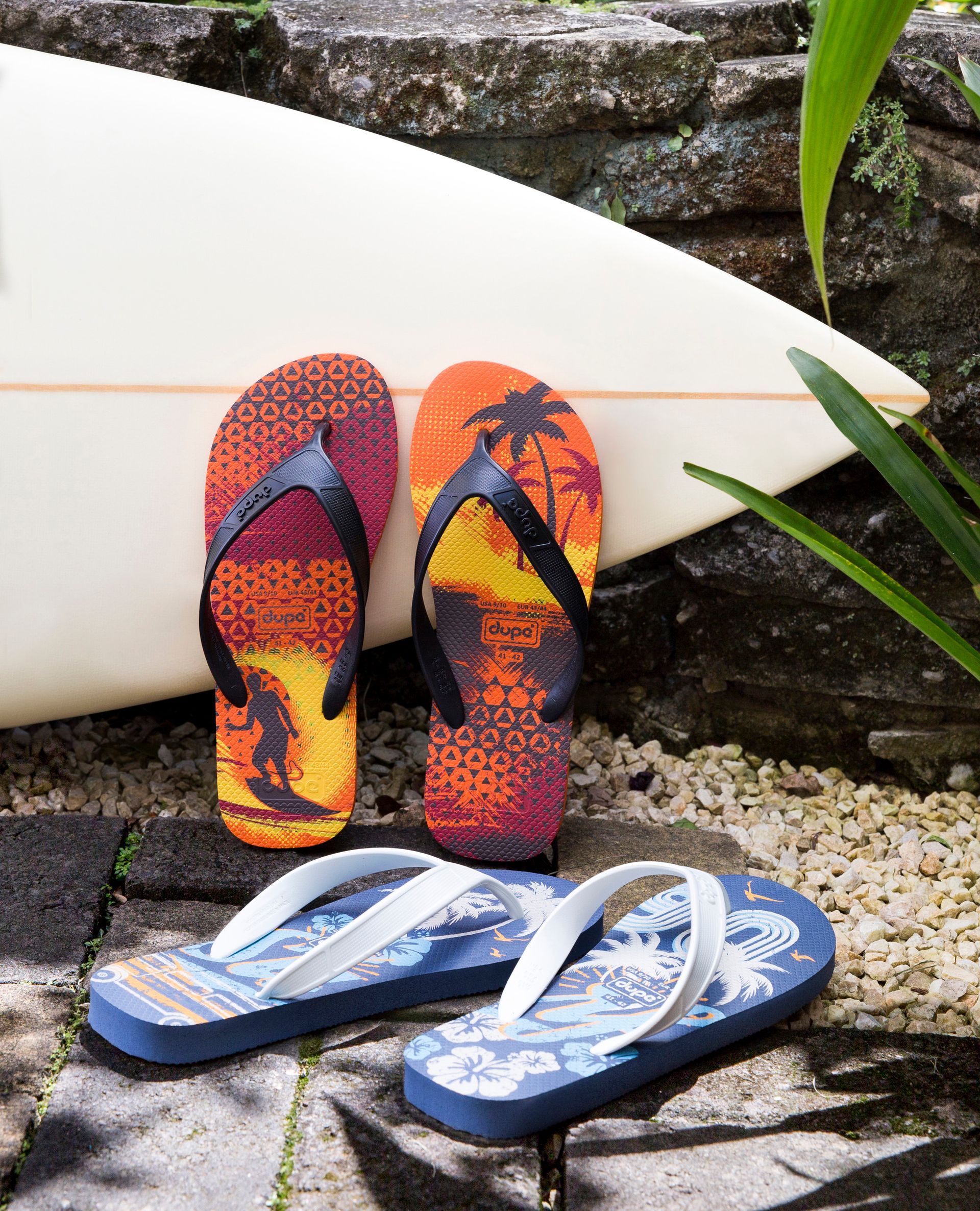 Duas chinelas coloridas apoiadas em uma prancha de surfe branca, com chinelas azuis sobre pedras à frente.