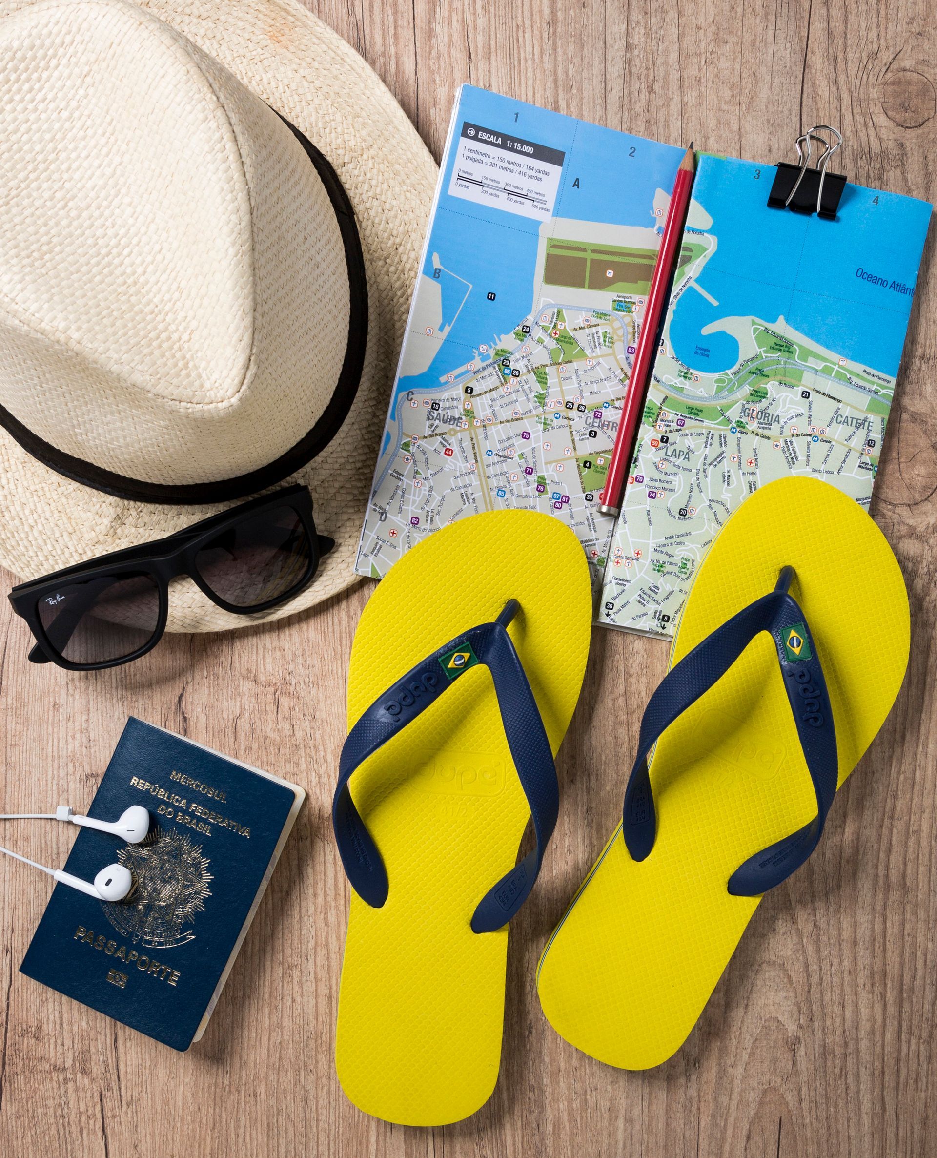 Itens essenciais para a praia: chapéu de palha, óculos de sol, chinelos, passaporte e mapas de viagem sobre uma superfície de madeira.