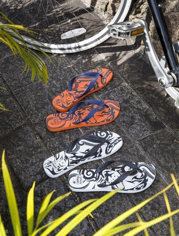 Chinelo de dedo com estampa laranja e preta sobre chinelo de dedo com estampa preta e branca em pavimento de pedra perto de rodas de bicicleta