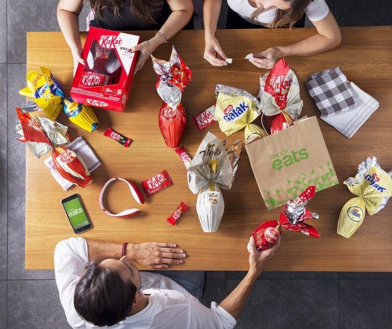 Pessoas ao redor de uma mesa compartilhando diversos lanches e bebidas embalados, em uma vista aérea.