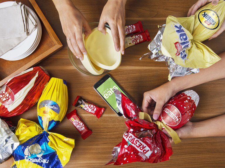 Mãos organizando doces coloridos embalados em uma mesa de madeira, com uma tigela no centro.
