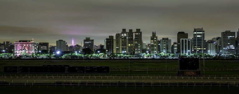Vista noturna da cidade com arranha-céus iluminados acima de uma rodovia ou ferrovia escura.