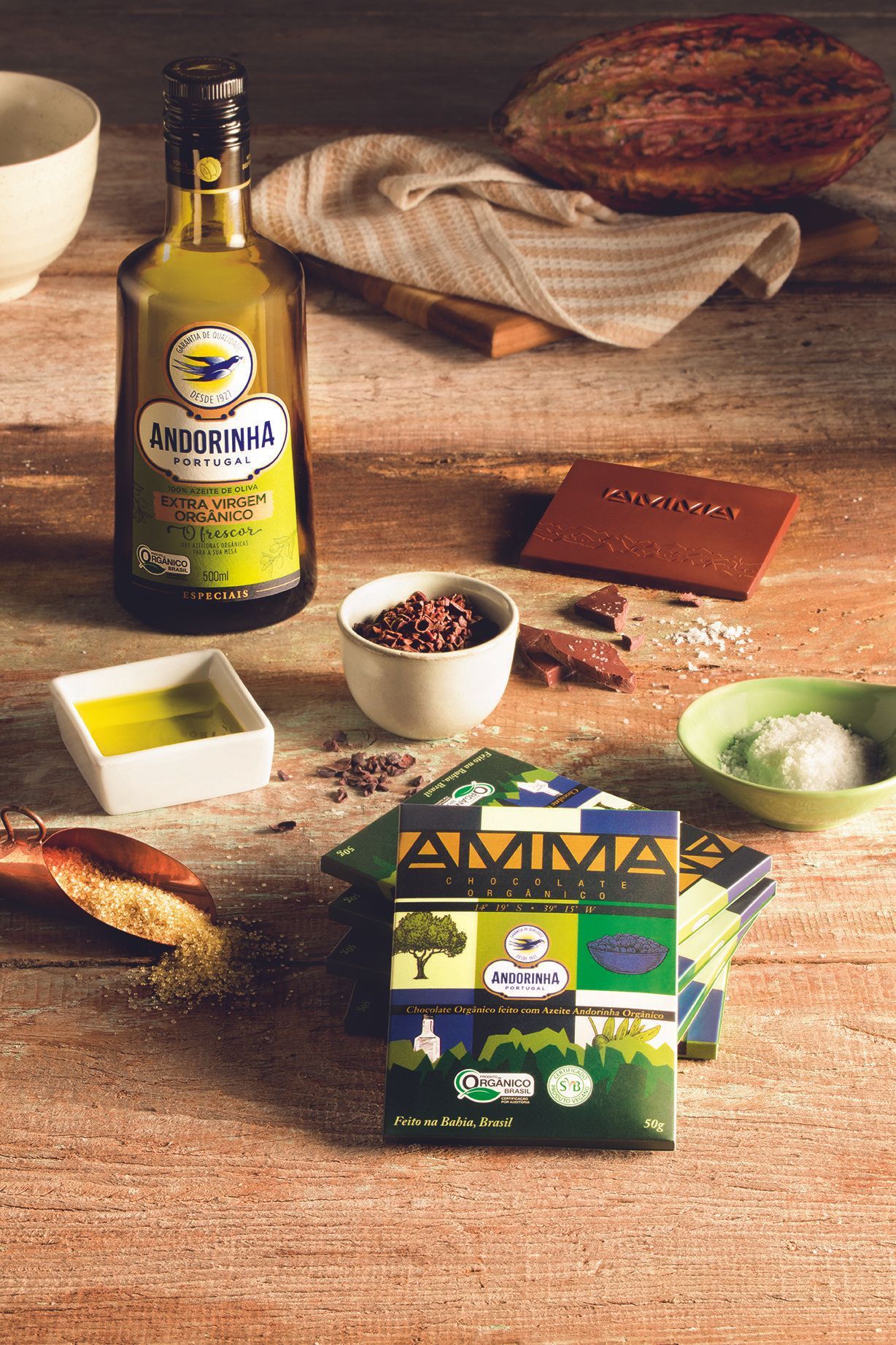 Garrafa de azeite com ingredientes e especiarias dispostas sobre uma mesa de cozinha rústica ao lado de uma caixa de produto.