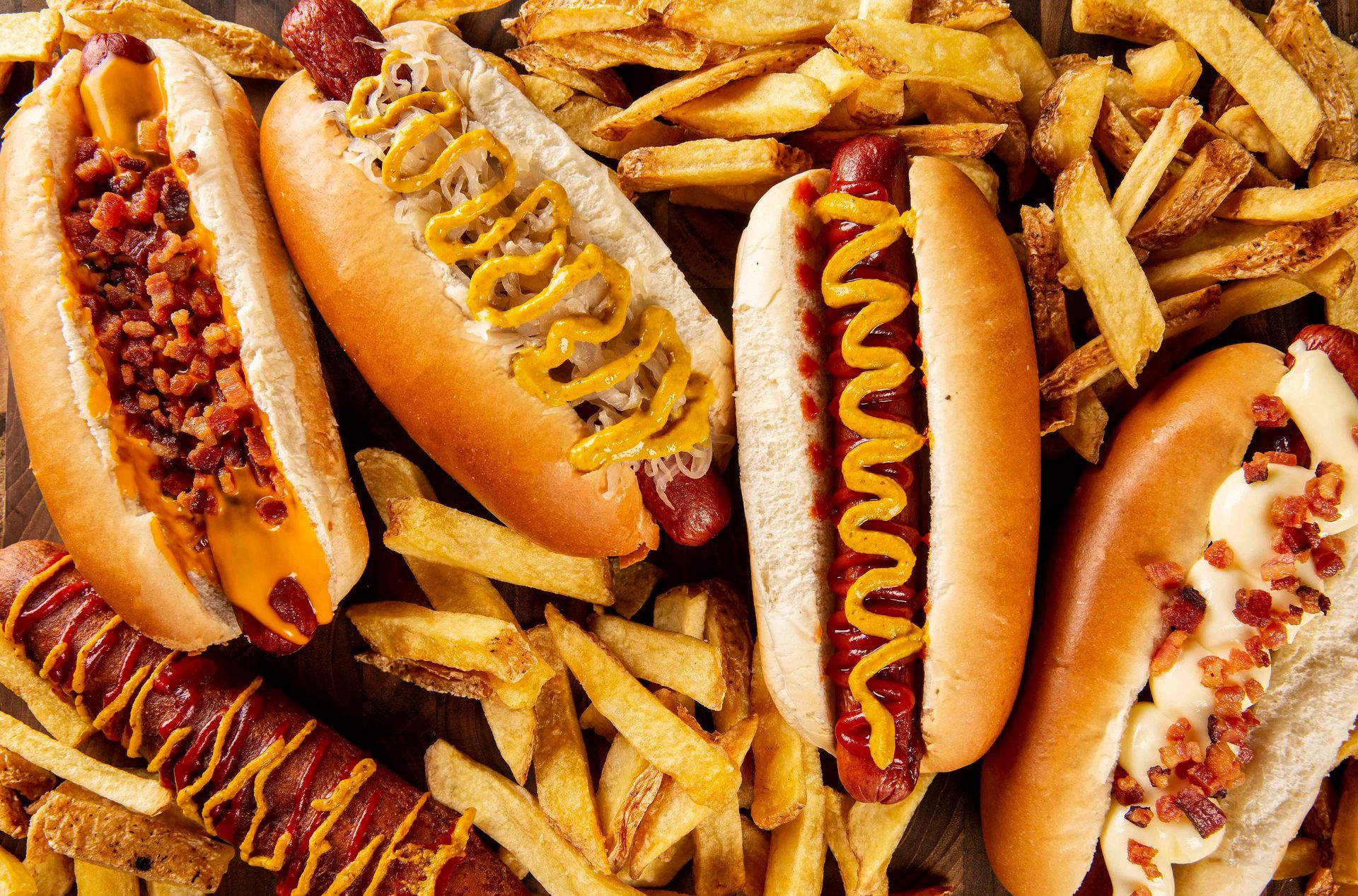 Cachorros-quentes variados cobertos com mostarda e ketchup, acompanhados de batatas fritas.