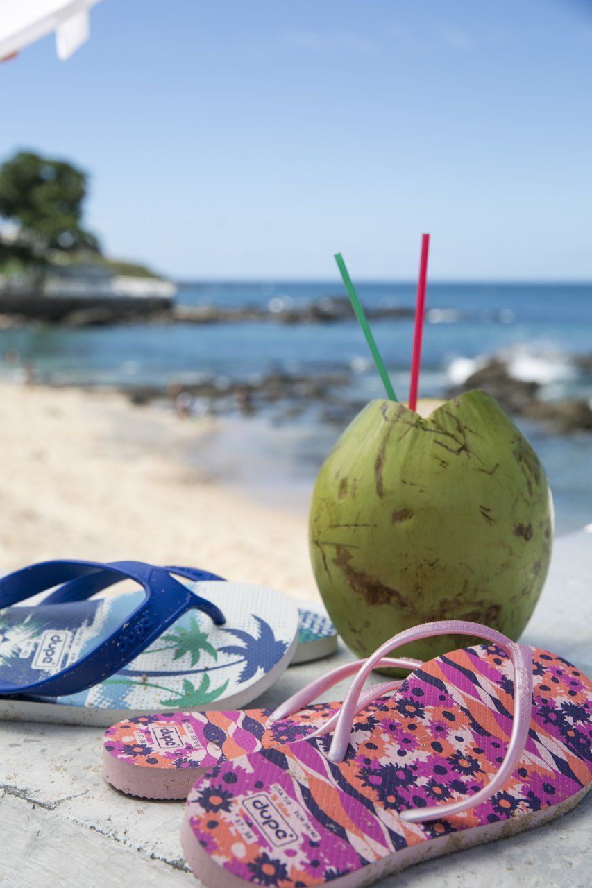 Bebida de coco com canudos e chinelos numa praia ensolarada à beira-mar.