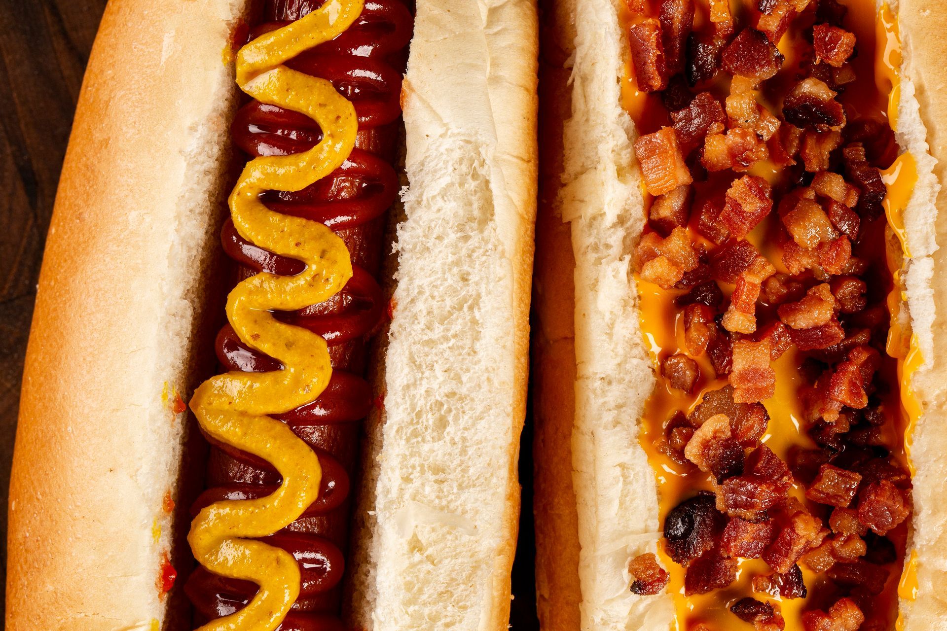 Close-up de dois cachorros-quentes, um com mostarda e outro com pedaços de bacon no pão.