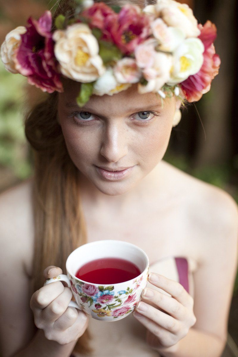 Mulher usando uma coroa de flores, segurando uma xícara de chá florida, olhando para a câmera.