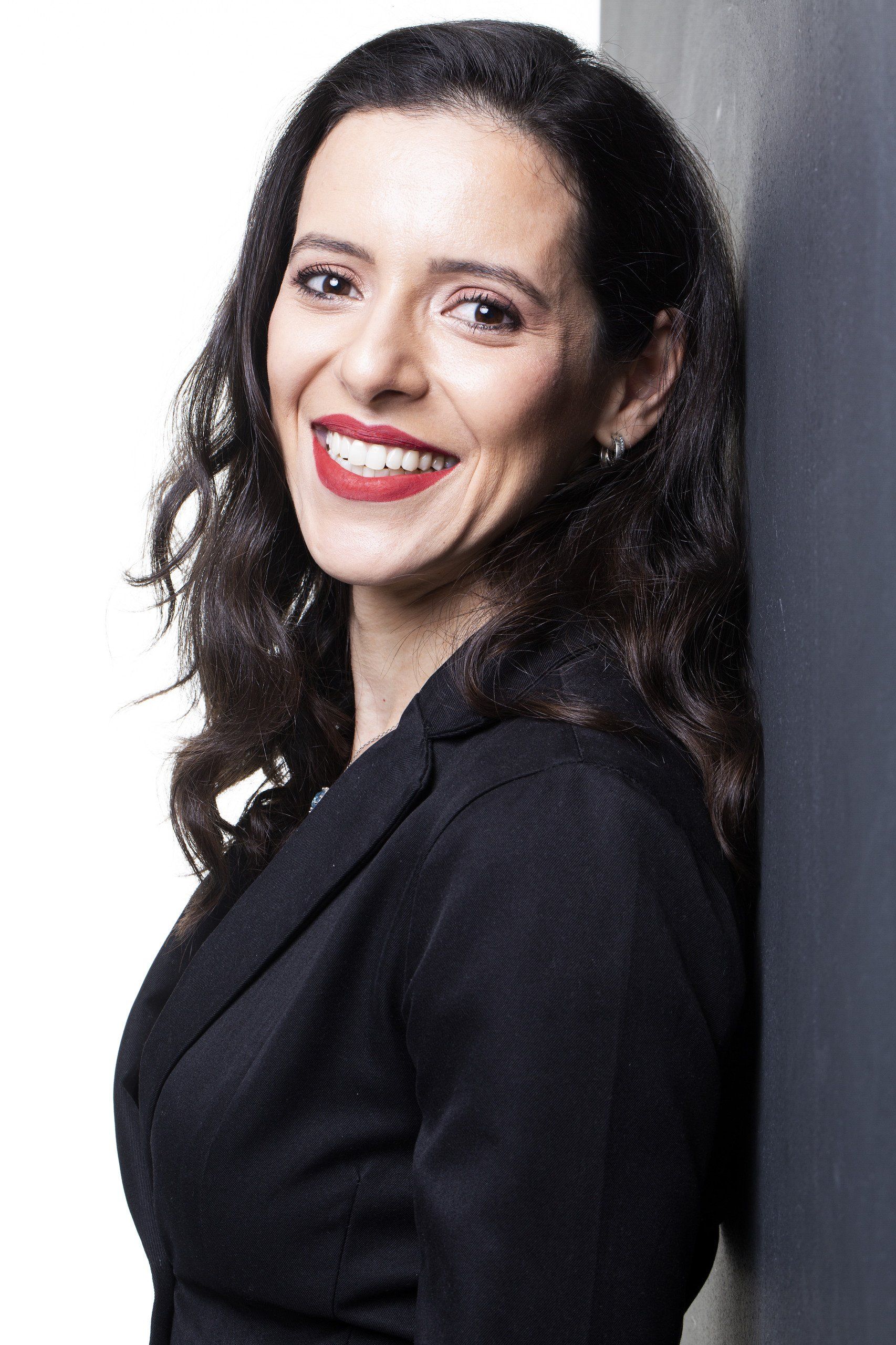 Mulher sorridente com cabelos escuros e cacheados, vestindo uma blusa preta, contra um fundo cinza claro.