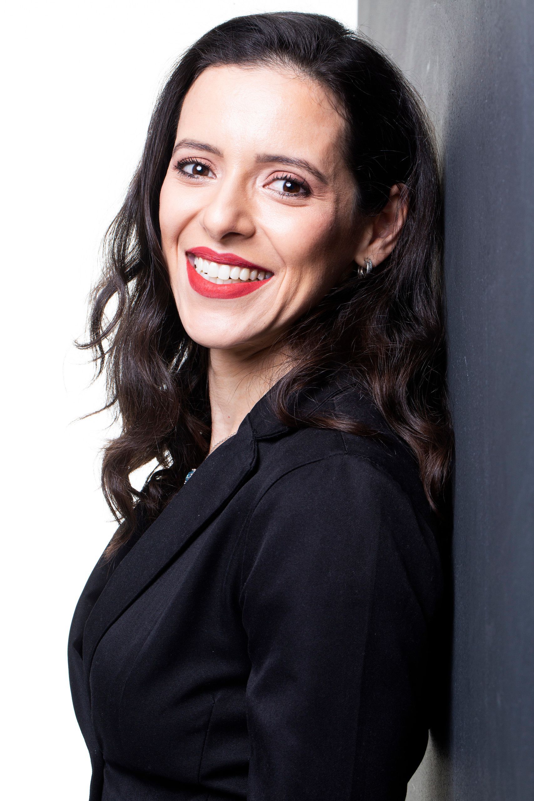 Retrato de uma mulher sorridente com cabelos escuros e cacheados, vestindo uma blusa preta contra um fundo cinza claro.
