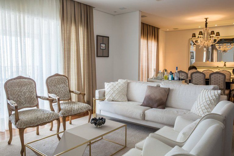 Sala de estar iluminada com sofá branco, duas cadeiras estampadas, mesa de centro de vidro e área de jantar ao fundo.