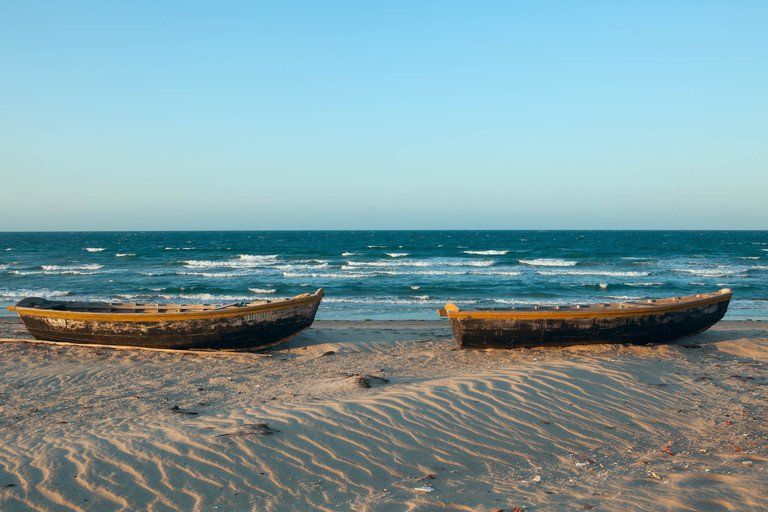 Dois barcos de madeira em uma praia de areia com o oceano e o céu azul ao fundo.