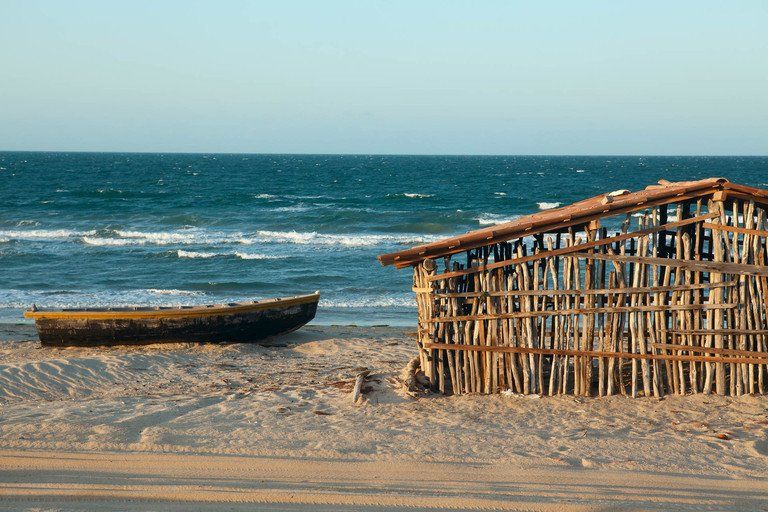Pequeno barco de madeira e cabana de pesca em uma praia de areia à beira-mar sob um céu claro.