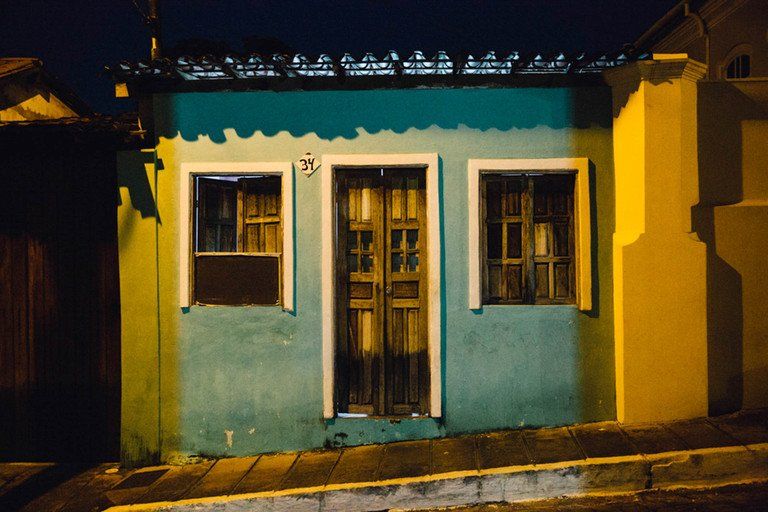 Pequena casa turquesa com porta amarela e janelas iluminadas à noite em uma rua tranquila.