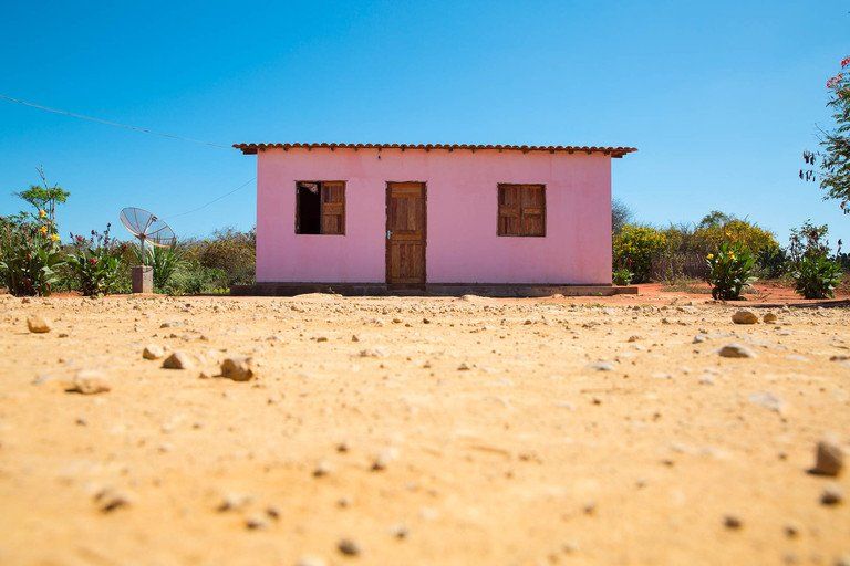 Pequena casa rosa com telhado vermelho em uma paisagem seca e arenosa sob um céu azul claro.