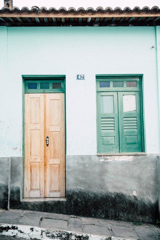 Duas portas em uma parede branca, uma bege e outra verde, com moldura inferior cinza desgastada.
