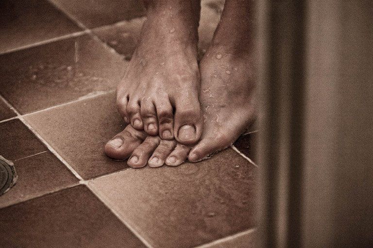 Pés descalços sobre um piso de azulejos, um pé sobre o outro, em um close-up com tons quentes de sépia.