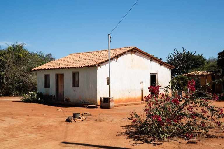 Pequena casa branca com telhado de telhas vermelhas em uma paisagem rural seca com arbustos e um poste de eletricidade.
