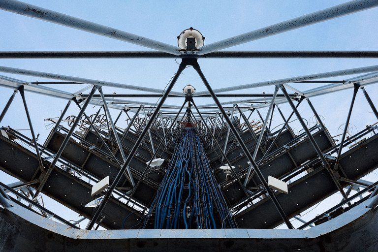 Vista interna de uma antena parabólica de radiotelescópio com suportes metálicos e um feixe de cabos azuis sob um céu nublado.