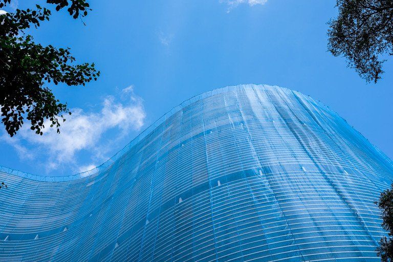 Arranha-céu de vidro azul com fachada curva contra um céu claro, emoldurado por galhos de árvores.