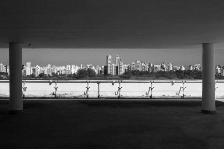 Vista em preto e branco do horizonte de uma cidade do outro lado da água, a partir de uma passarela coberta com colunas.
