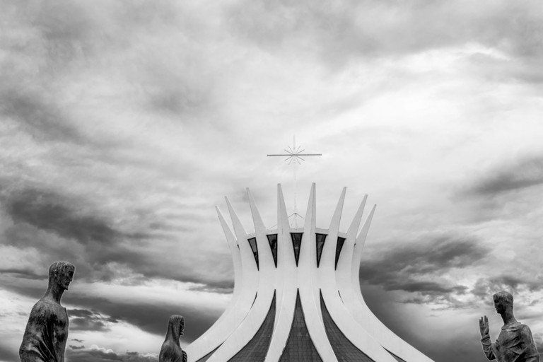 Escultura em preto e branco com pináculos curvados para cima contra um céu nublado, ladeada por duas estátuas.