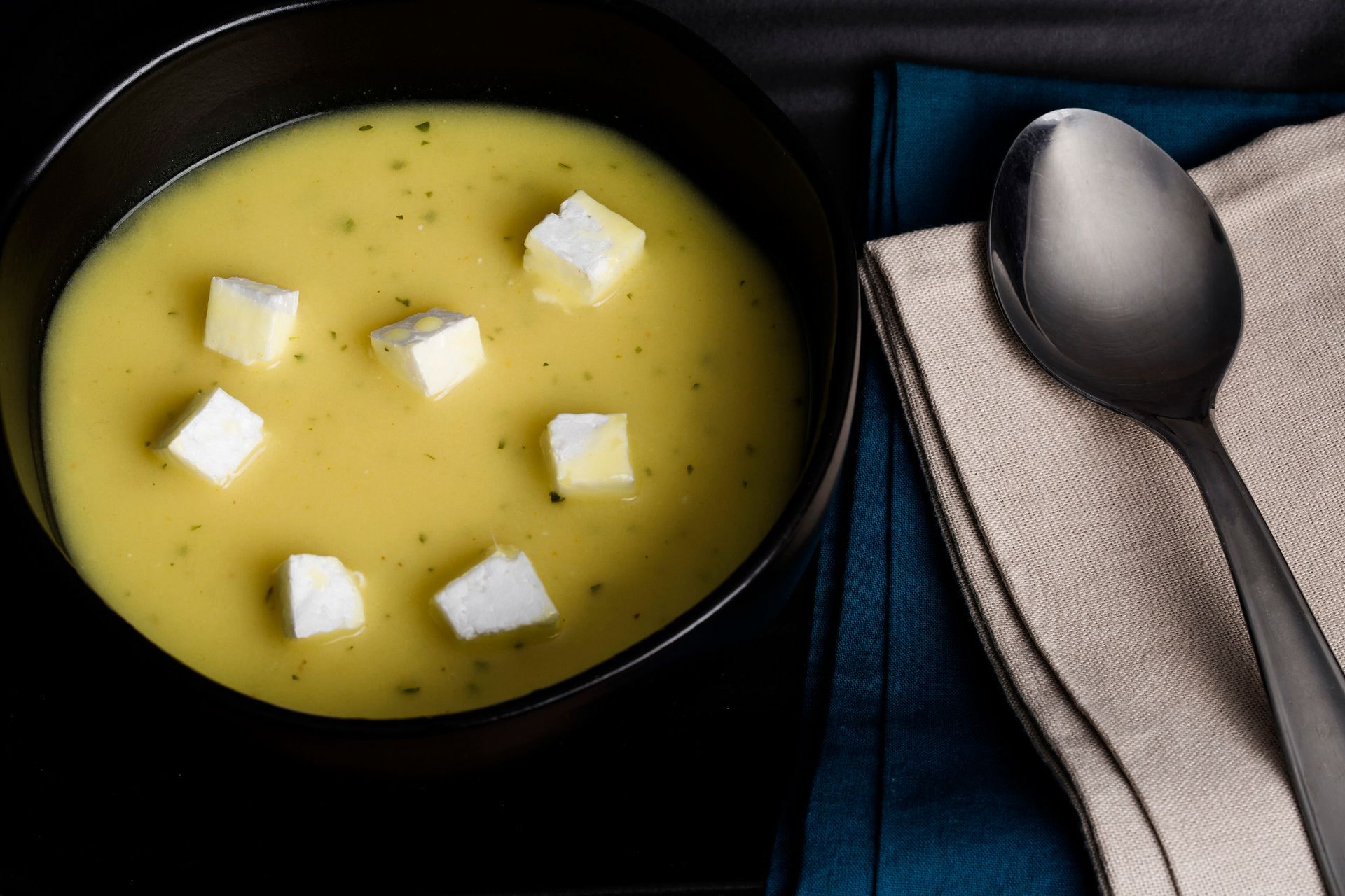 Tigela de sopa amarela com cubos de queijo branco ao lado de uma colher e um guardanapo sobre uma mesa escura.