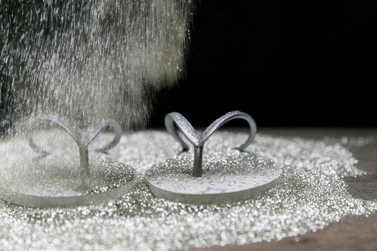 Close-up de duas argolas de metal puxando lacres sobre uma superfície prateada texturizada, com pó brilhante caindo de cima.