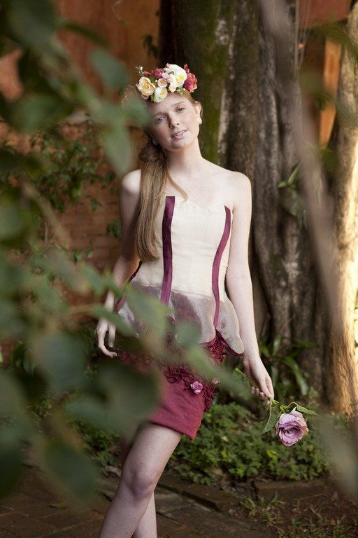 Mulher com vestido cor de vinho e coroa de flores posando ao ar livre entre árvores, segurando uma flor roxa.