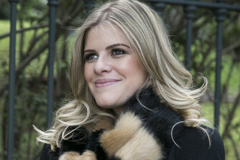 Mulher loira de casaco preto, sorrindo ao ar livre em frente a uma cerca.