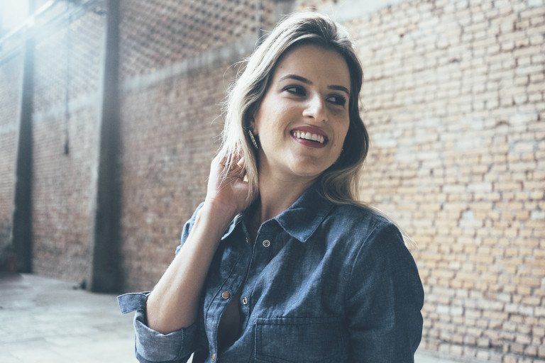 Mulher de camisa jeans sorrindo ao lado de uma parede de tijolos, com a luz do dia entrando suavemente pela esquerda.