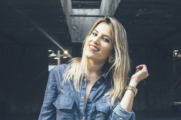 Mulher loira sorridente, vestindo camisa jeans, posando sob uma cobertura industrial.