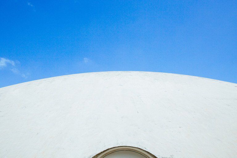 Cúpula branca curva contra um céu azul brilhante, com uma pequena janela circular na borda inferior.