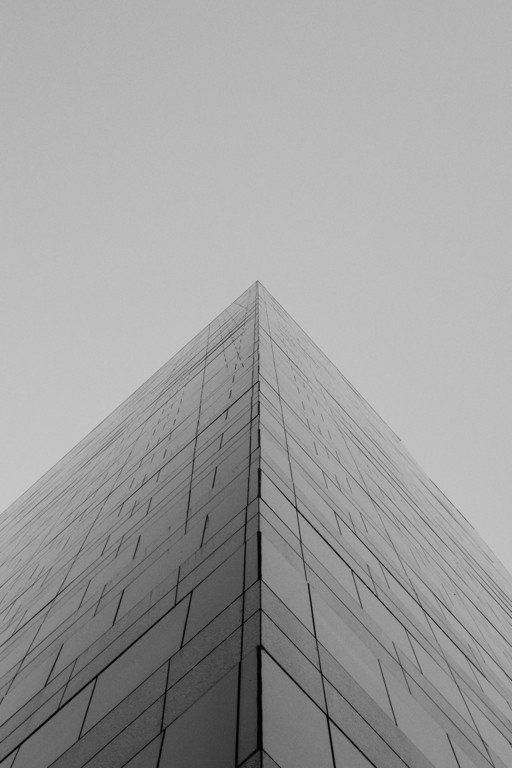 Vista em preto e branco da esquina de um alto edifício de vidro que se ergue contra um céu nublado.