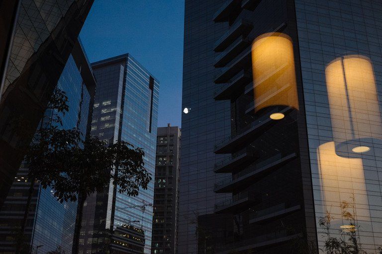 Vista da cidade ao entardecer com torres de escritórios de vidro e luzes cilíndricas brilhantes.