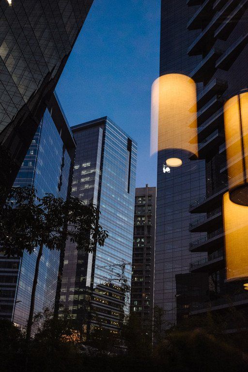 Rua da cidade ao entardecer, entre altos edifícios de vidro, com lâmpadas de luz quente em primeiro plano.